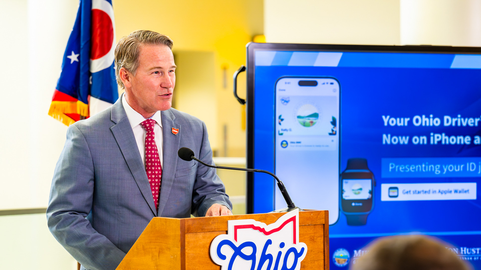 Senator Jon Husted speaking at a podium in front of the Ohio flag