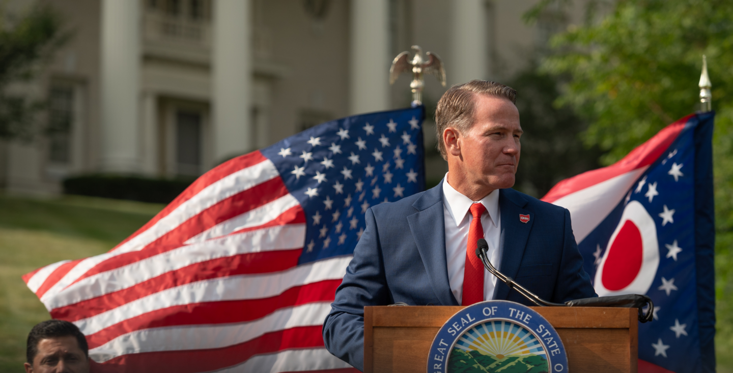 Senator Jon Husted speaking at a podium in front of the US and Ohio flags