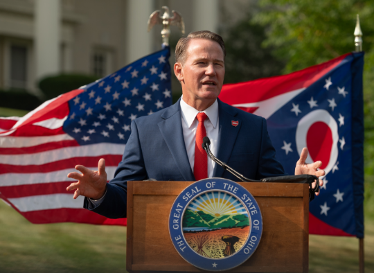 Senator Jon Husted speaking at a podium in front of the US and Ohio flags