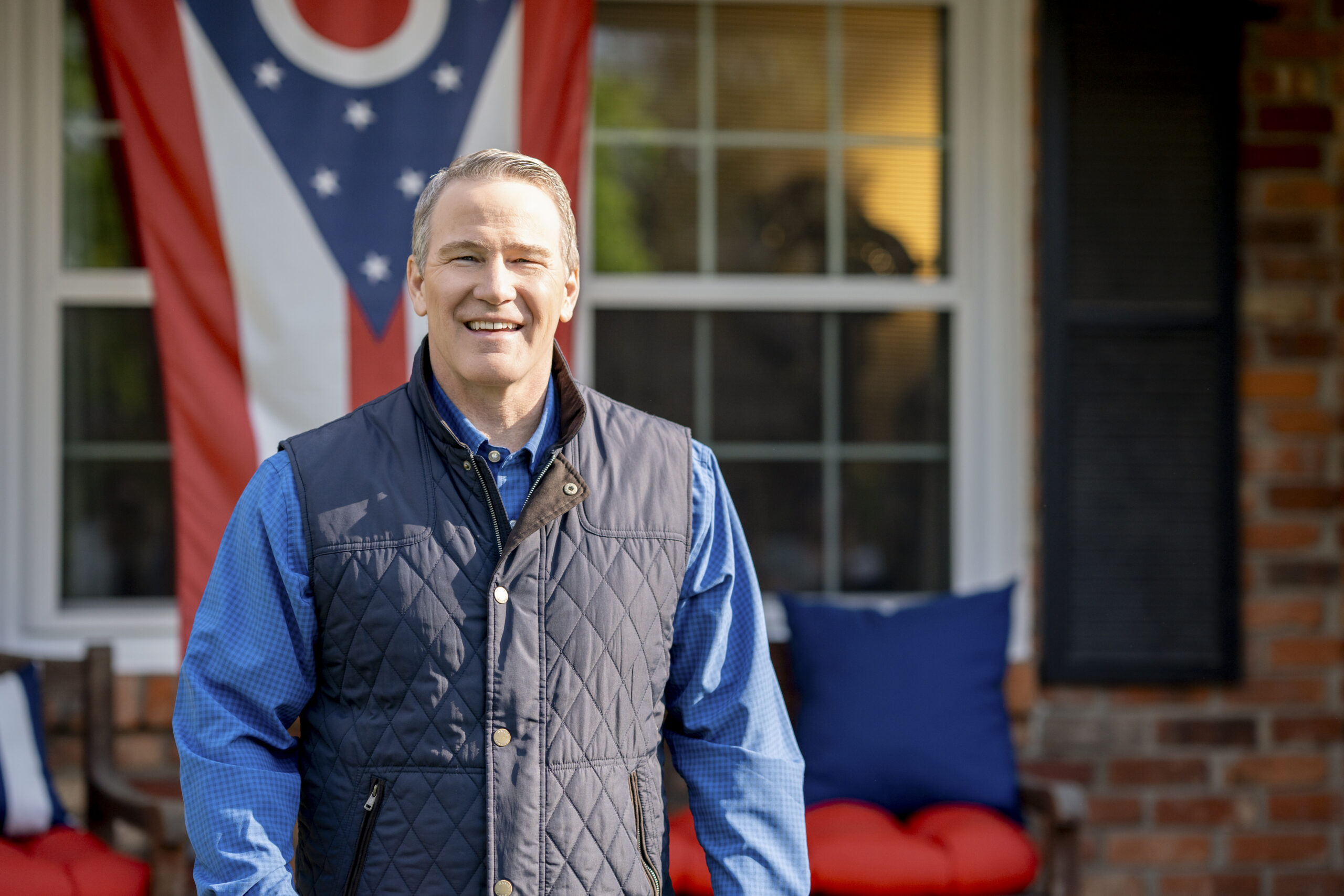 Jon Husted in a blue shirt and gray quilted vest standing outside a brick house with a patriotic flag in the background.
