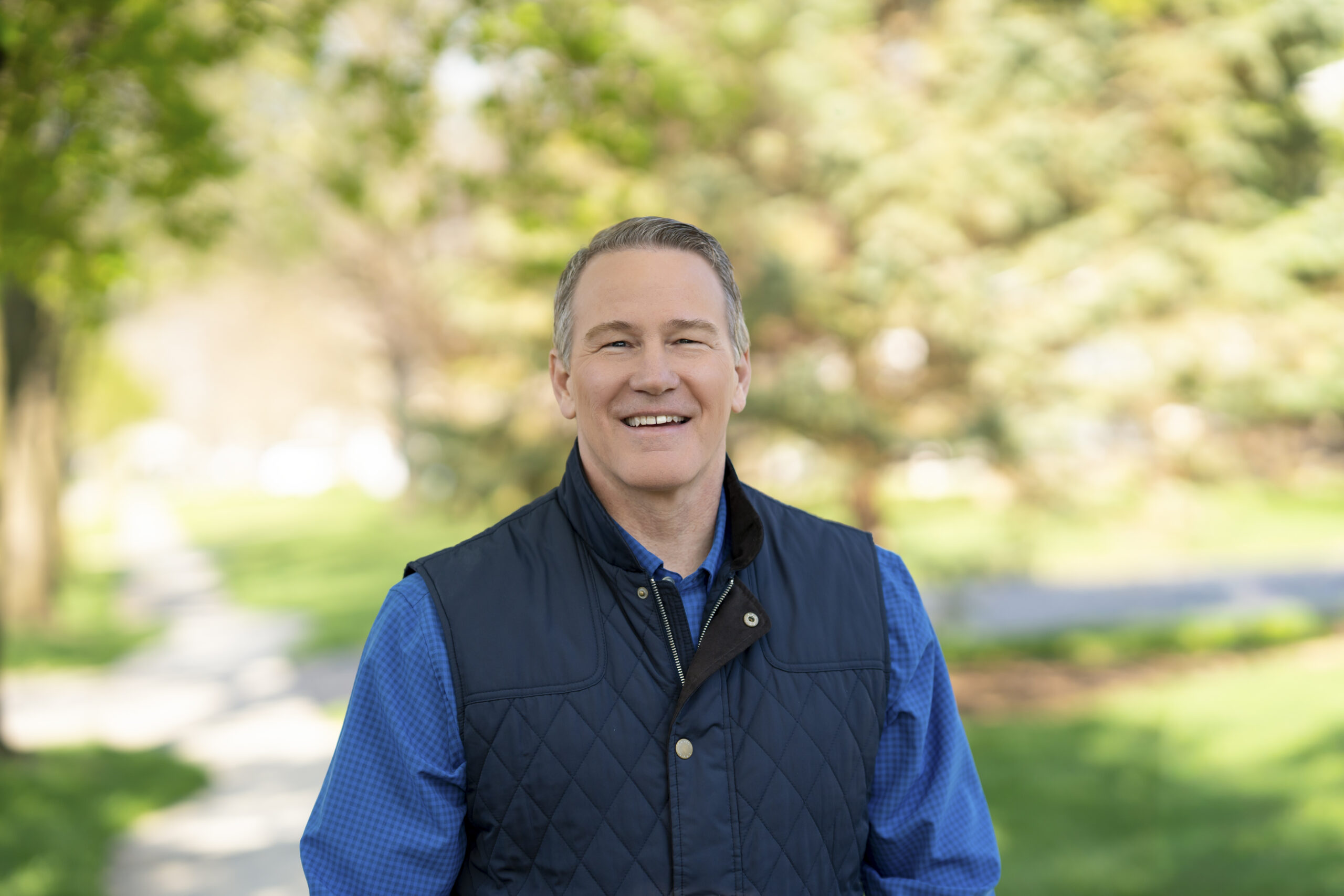 Jon Husted in a blue shirt and navy quilted vest standing in a sunlit park with greenery in the background, facing the camera.