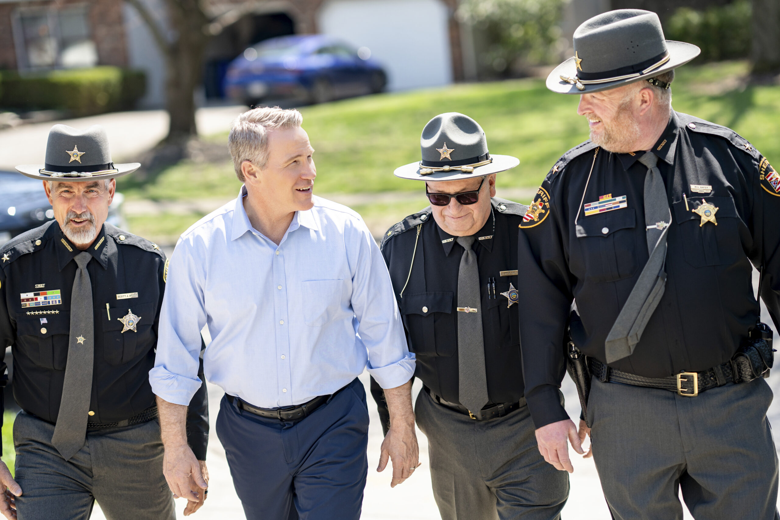Jon Husted in a light blue shirt walking and smiling with three uniformed police officers outdoors on a sunny day, as they chat together.