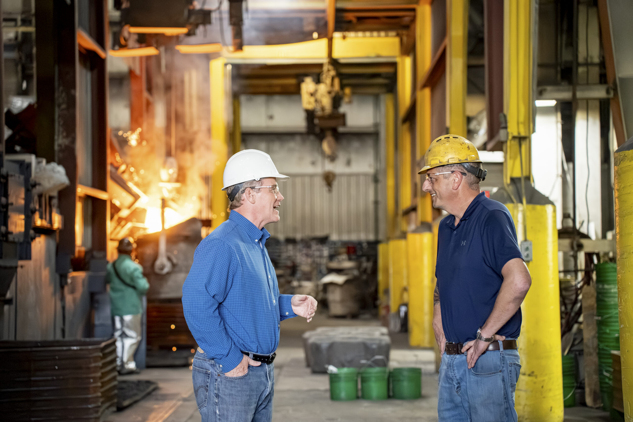 Jon Husted talks with factory worker and smile in a busy industrial facility with yellow support beams behind them, safety gear on.