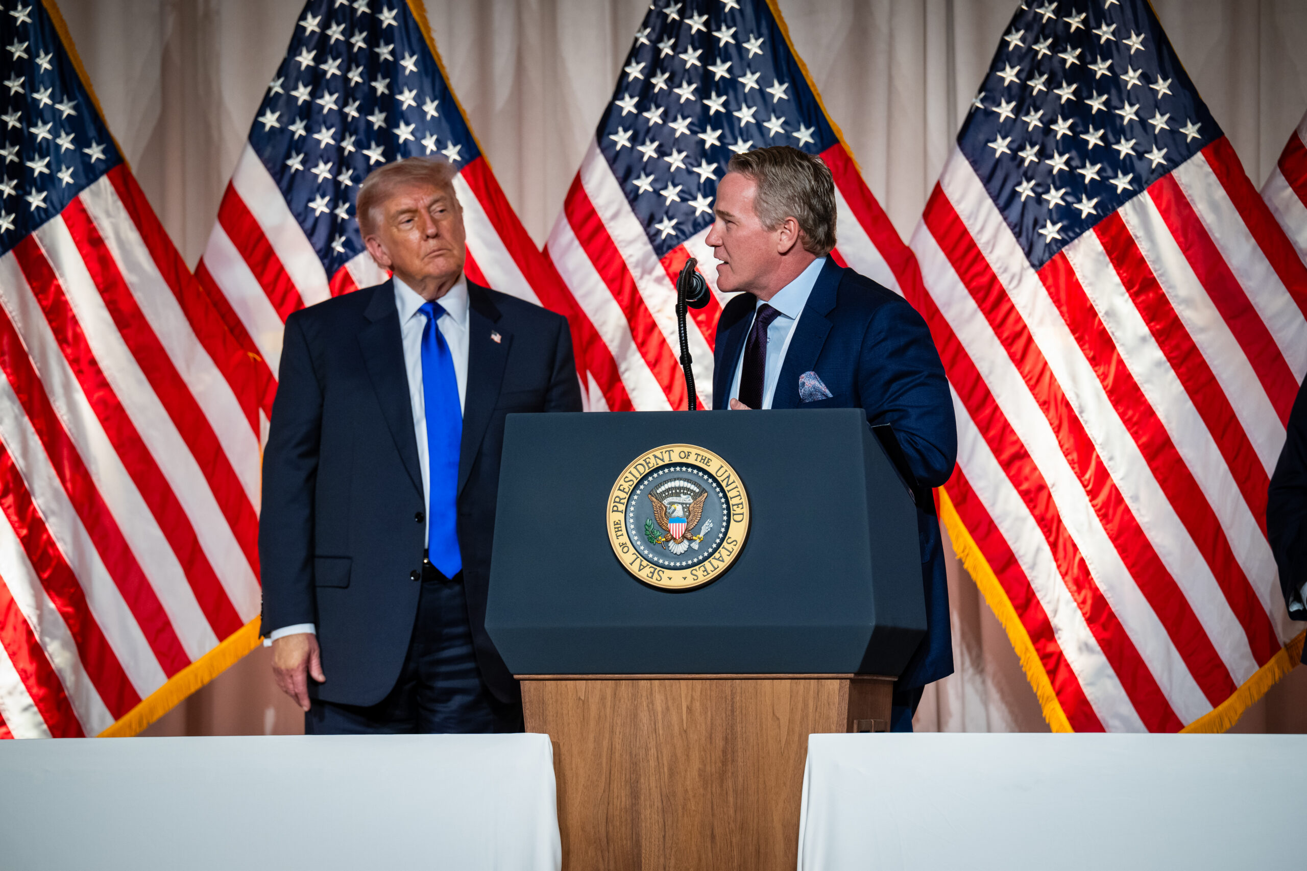 Two men stand at a podium with the presidential seal, flanked by multiple American flags as they speak.