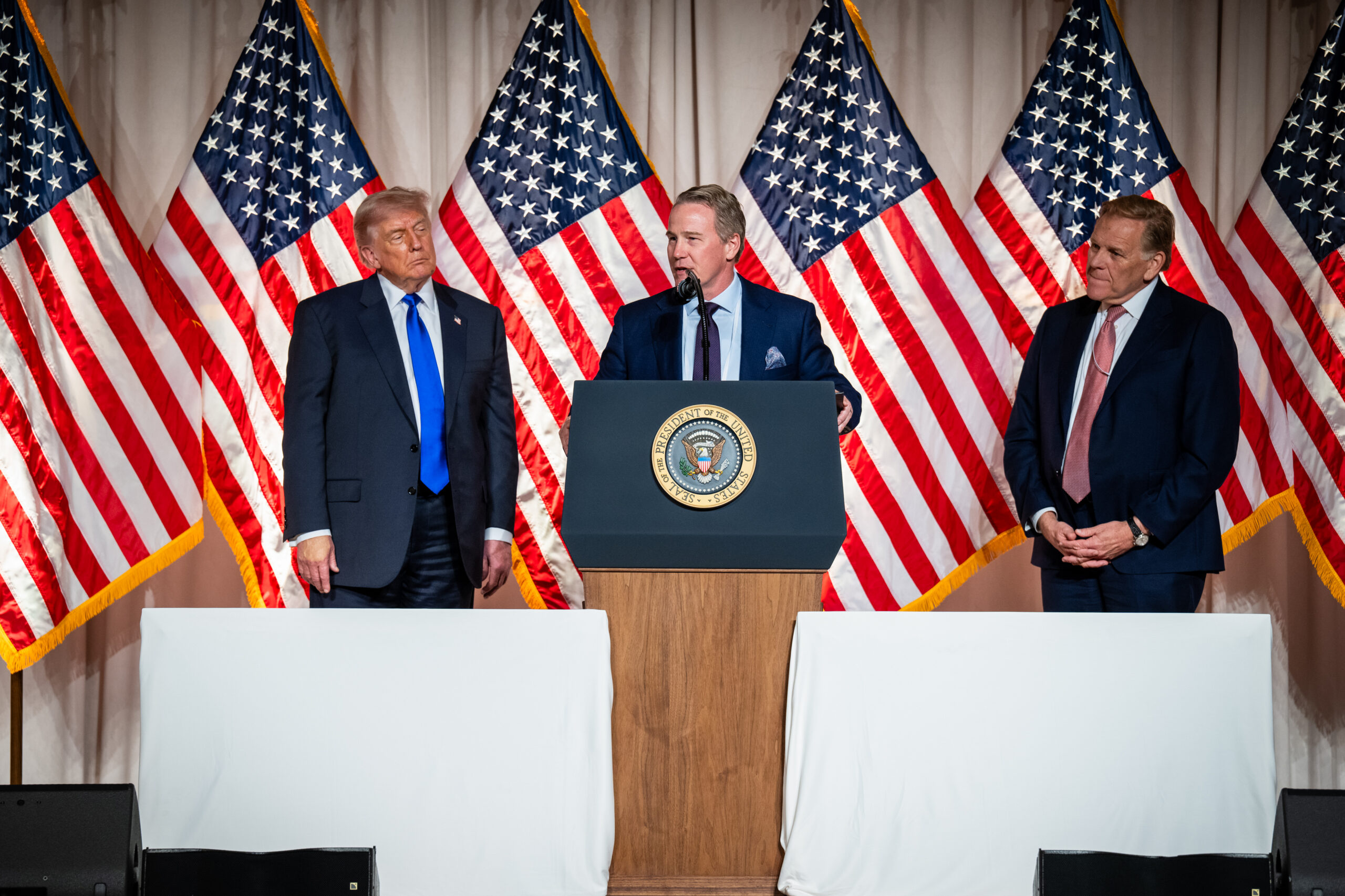 Three men in suits on stage with U.S. flags behind; central speaker at a lectern with the presidential sealed badge.