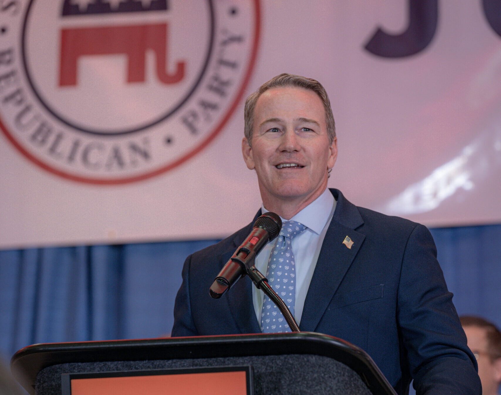 Man in a dark suit speaks at a podium with a microphone, a floral arrangement nearby and a large Republican Party seal in the background.