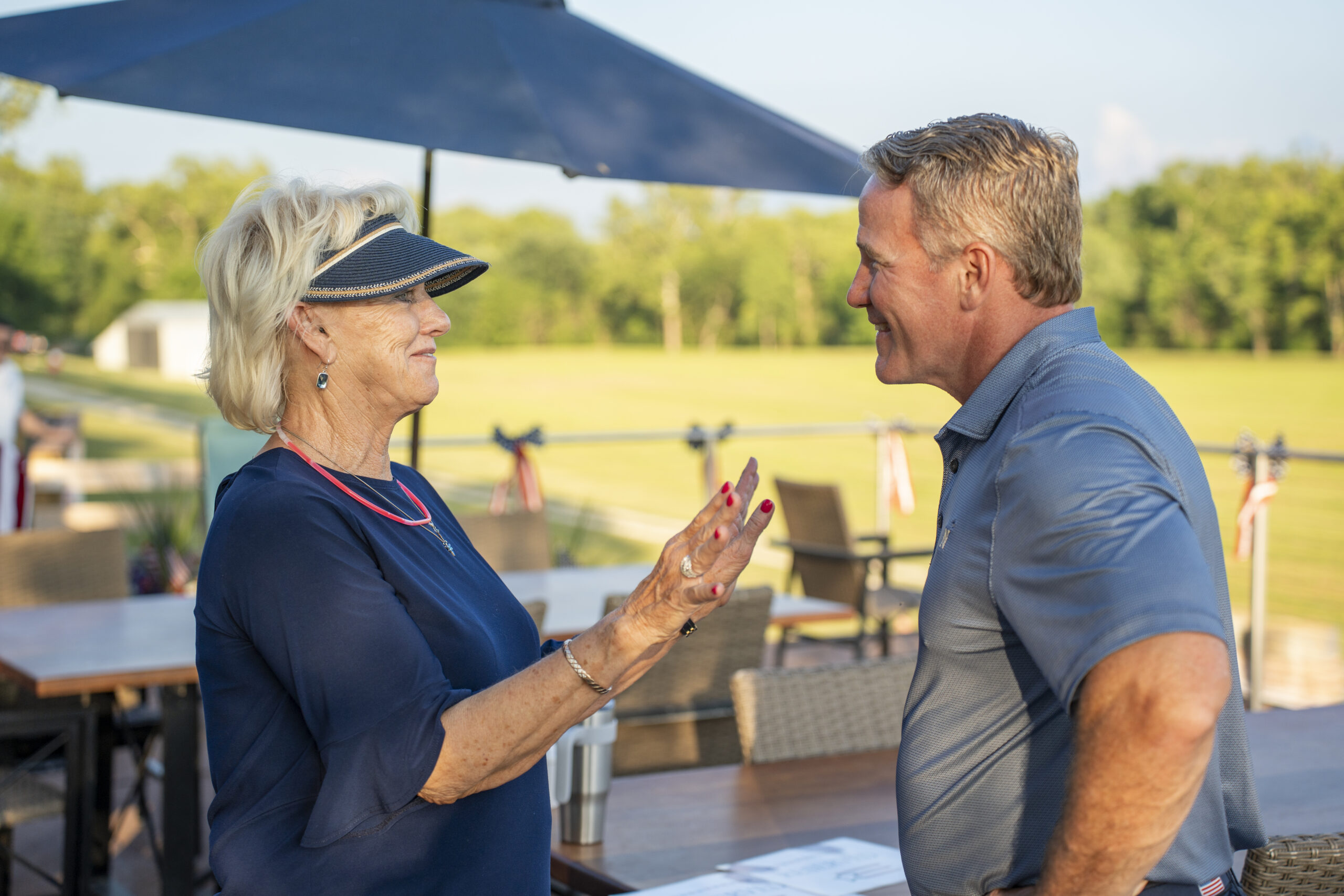 Two adults converse outdoors at a cafe, facing each other with hands gesturing as they talk under an umbrella.