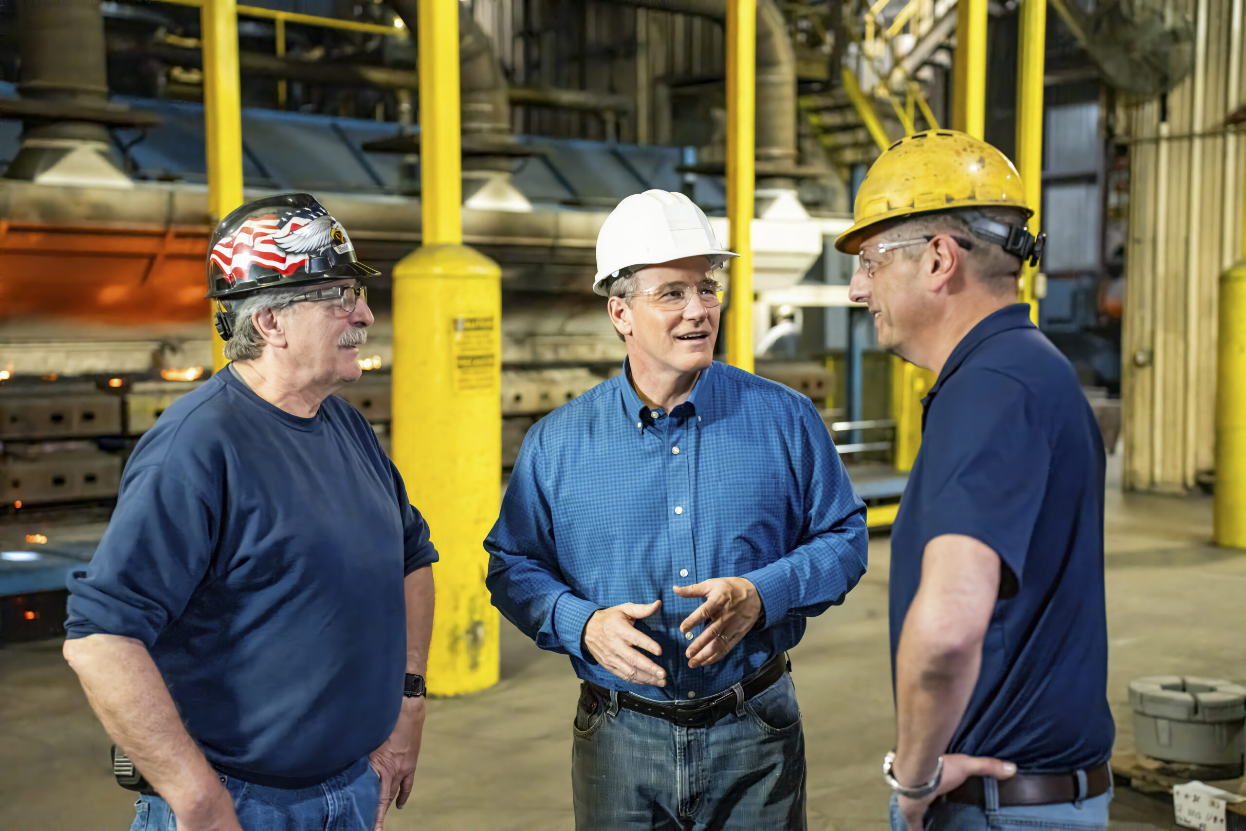 Three workers wearing hard hats and safety glasses converse in an industrial plant with yellow safety posts in the background.