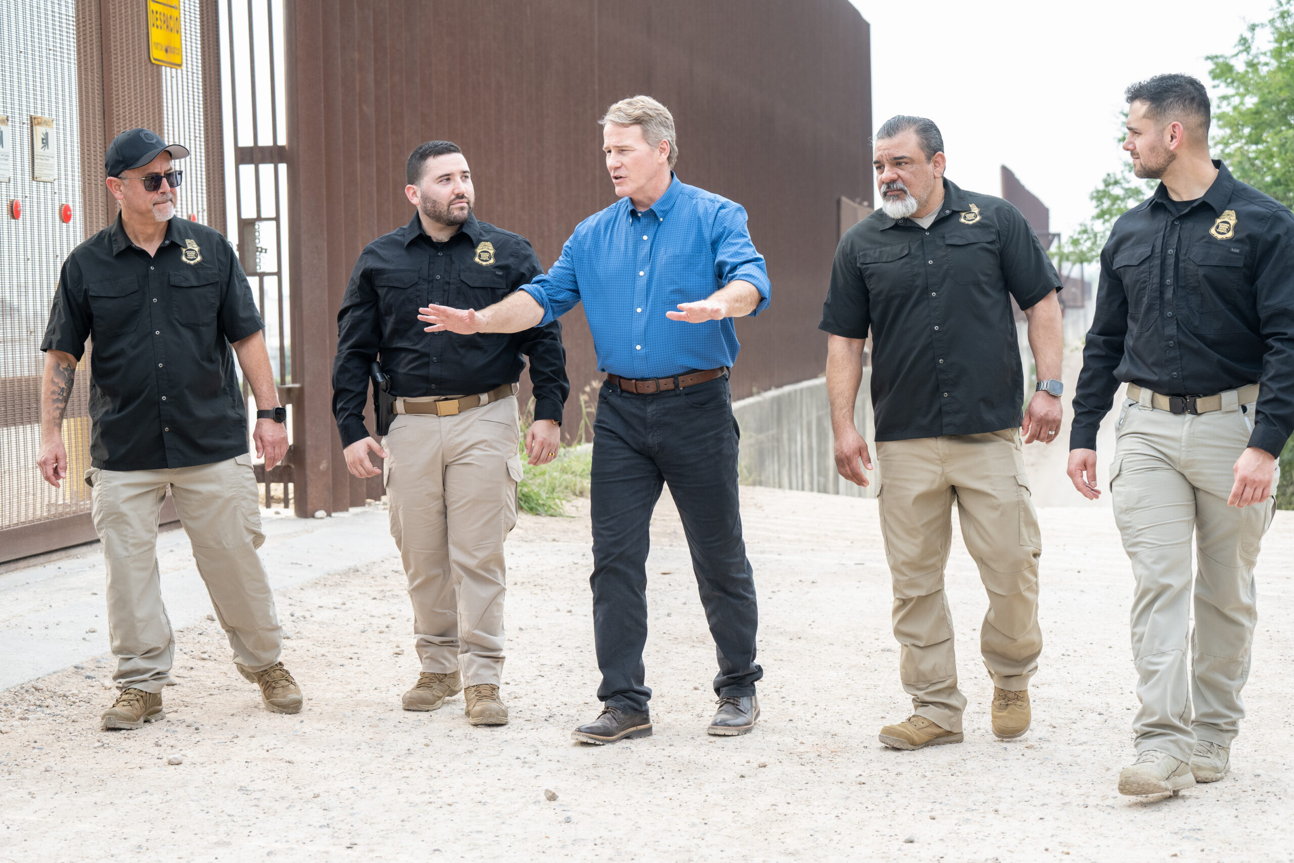 Group of five men walking along a dusty outdoor path; four wear black uniforms with badges, one in a blue shirt gesturing as if leading a discussion near a tall brown fence.