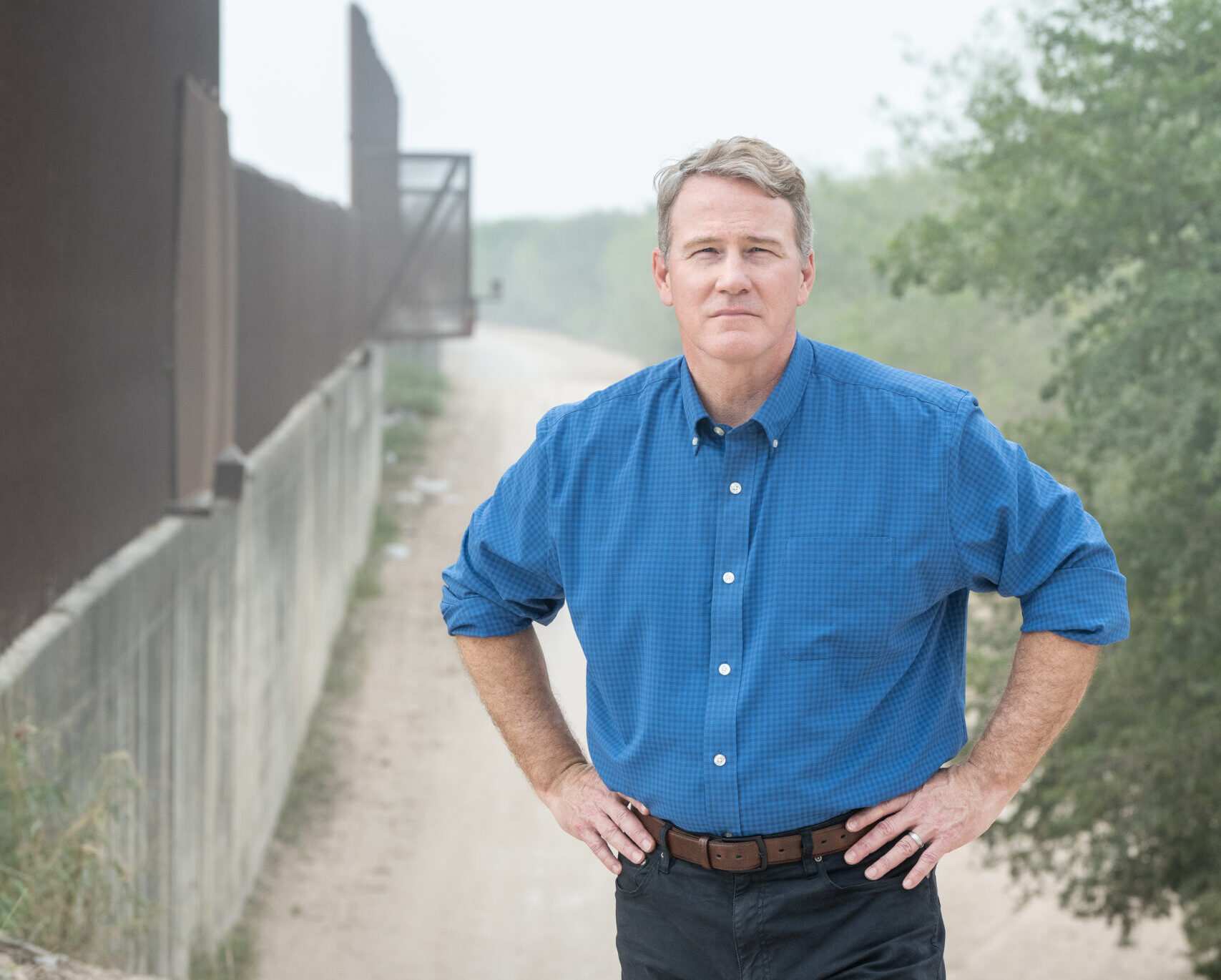 Jon Husted in a blue checkered shirt stands with hands on hips on a dirt path beside a tall fence outdoors.